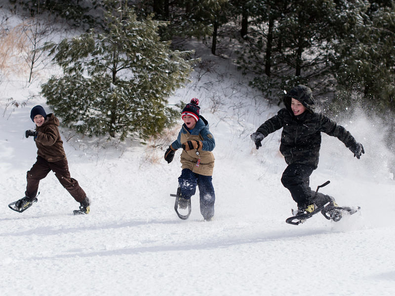 A group of kids walks through a snowy field on snowshoes, with woods in the background. (photo © Ben Conant)