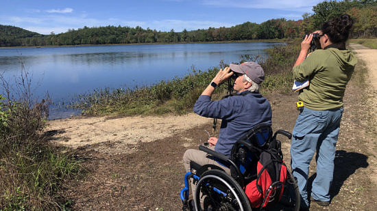 Two people -- one standing, one in a wheelchair -- hold binoculars up to their eyes while birding at a large lake. (photo © Jim Hassinger)