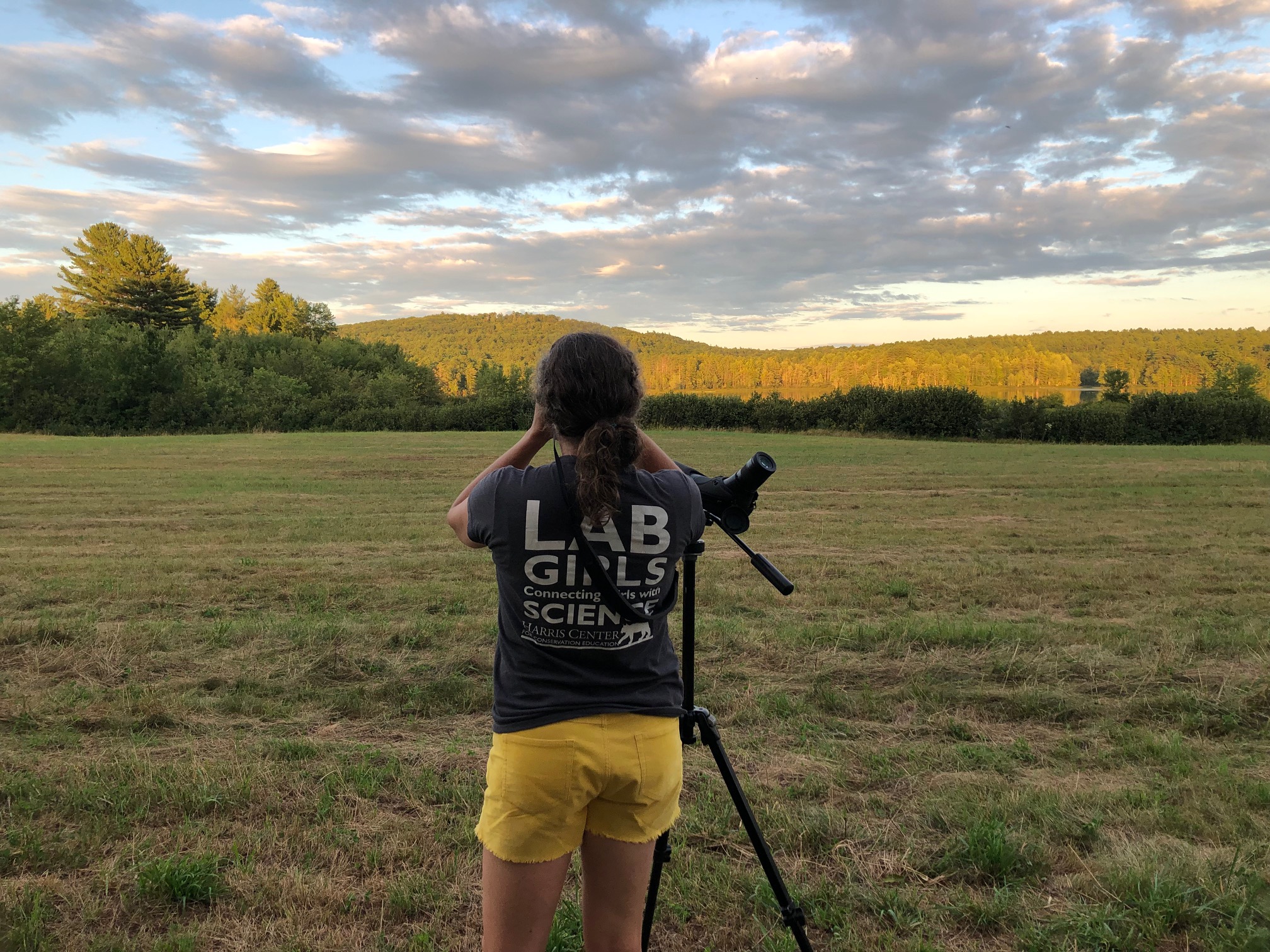 A woman stands in a field, looking to the distance. She is wearing a shirt that says "LAB GIRLS: Connecting Girls with Science" and standing in front of a spotting scope. The hills in the distance glow in late-day sun. (photo © Phil Brown)