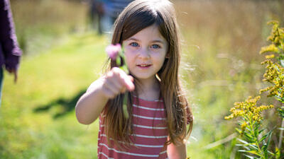 A young girl holds a small flower while standing in a sunny field. (photo © Ben Conant)