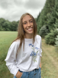 Carissa Miller smiles while standing in a mowed field lined with trees. (photo courtesy of Carissa Miller)